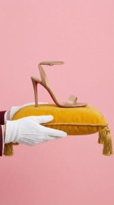 Paire de talons aiguilles beiges Louboutin présentées sur un coussin en velours jaune par des mains gantées. Exemple de direction artistique et de captation publicitaire en studio par Noswilprod.